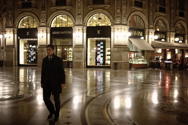 Homme se promenant dans le quartier de la mode de Milan en soirée. Derrière lui, les magasins de luxe Louis Vuitton et Savini.