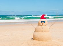 Un bonhomme de neige en sable sur une plage devant une mer verte. Top 25 des destinations Noël.