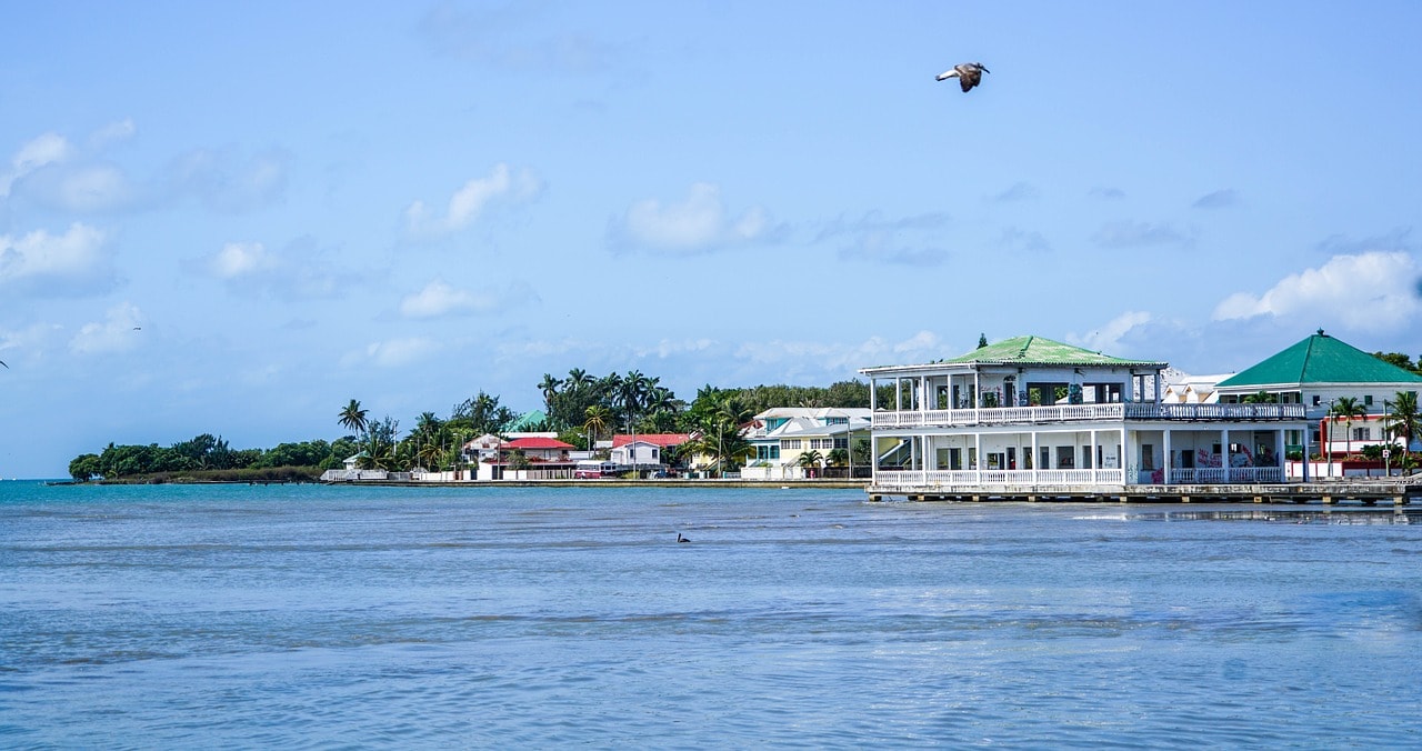 Un vol Belize pas cher avec Algofly illustré par des habitations en front de mer sous un ciel bleu.