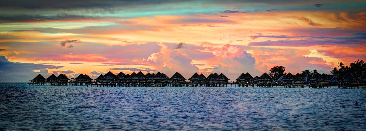 Un vol Bora Bora pas cher avec Algofly illustré par des maisons sur pilotis devant un somptueux coucher de soleil.