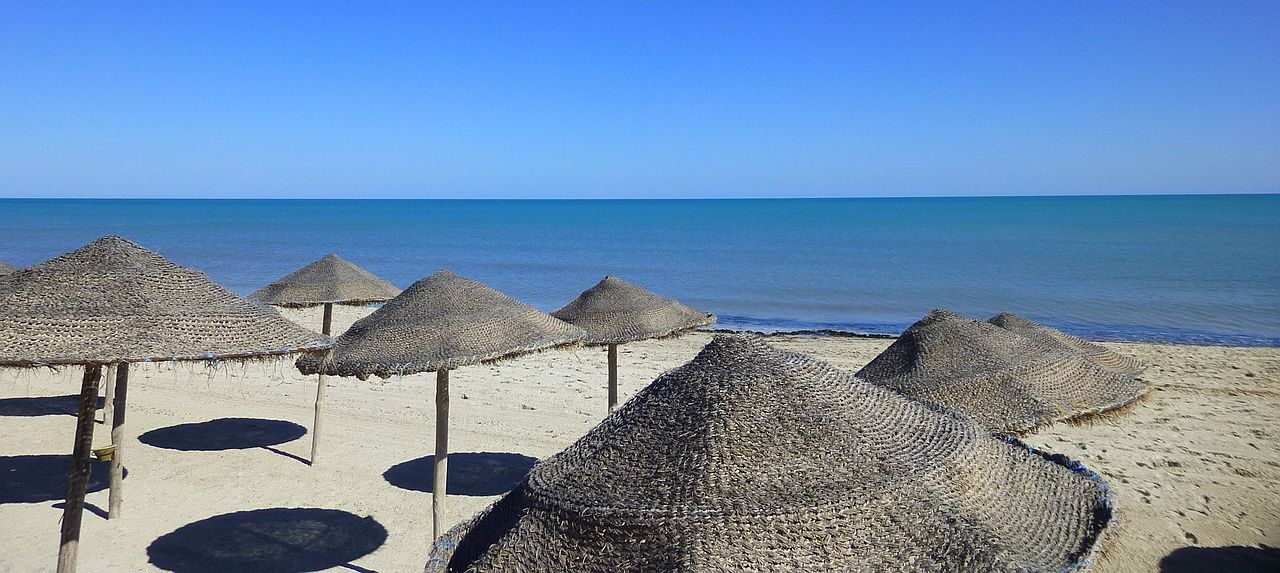 Un vol Djerba pas cher avec Algofly illustré par des parasols de jonc naturel sur une plage de sable blanc.