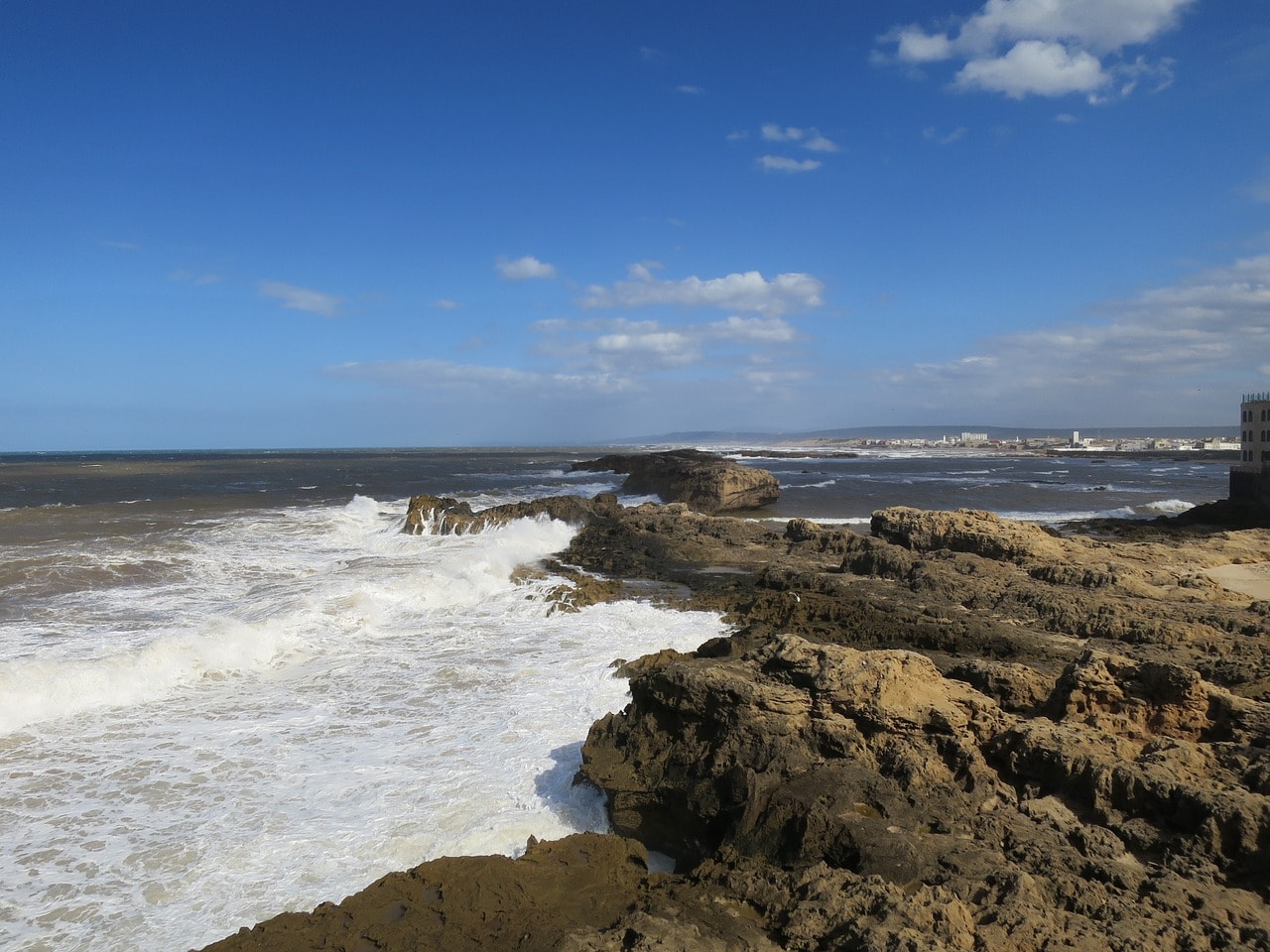 Un vol Essaouira pas cher avec Algofly illustré par une mer d'écume fouettant des rochers.