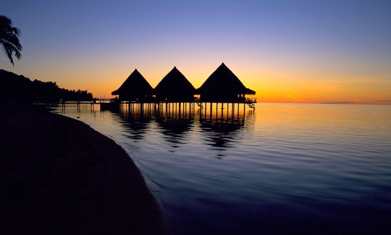 Un vol Tahiti pas cher avec Algofly illustré par un soleil couchant derrière les typiques maisons sur pilotis. 